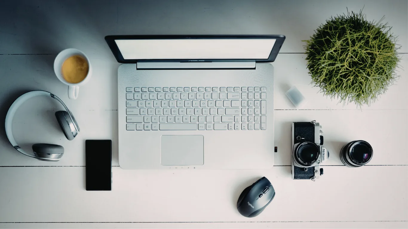 Top-down view of a laptop on a white desk with coffee, headphones, smartphone, mouse, camera and plant nearby.