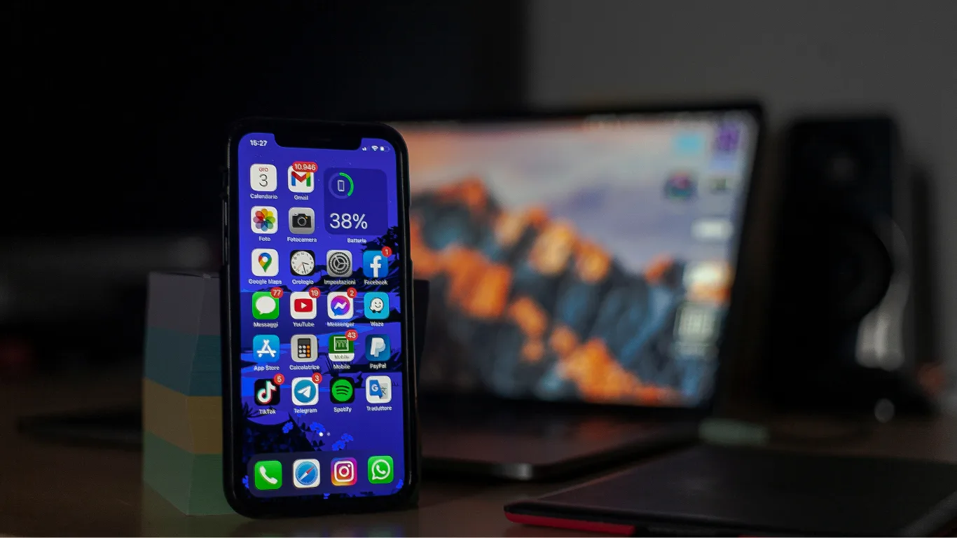 Smartphone with app icons on the home screen on a desk, a blurred laptop in the background.