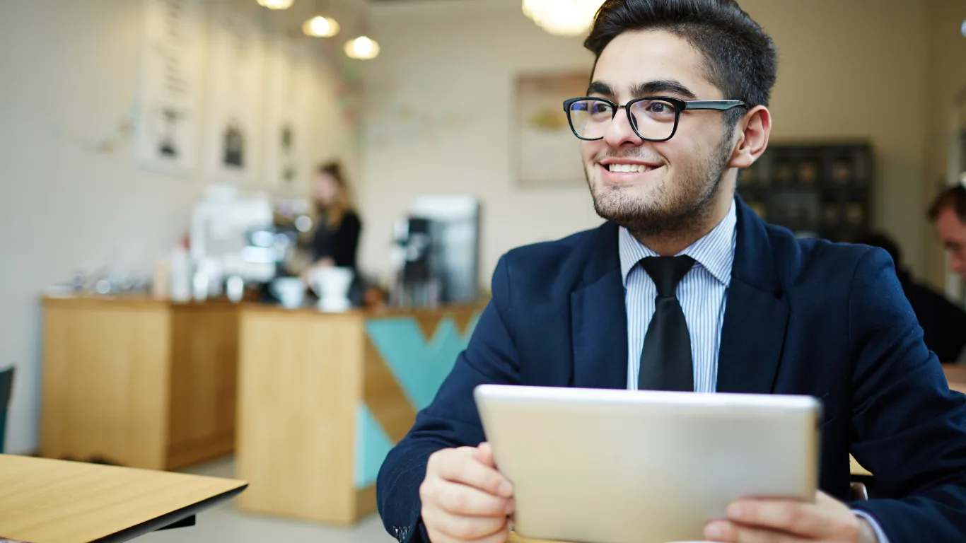 Smiling man in glasses and a navy blazer sits in a cafe, holding a tablet.