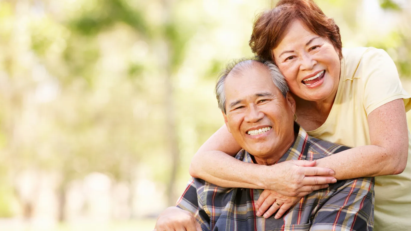 Smiling elderly couple in a sunlit park, the woman hugging the man from behind.