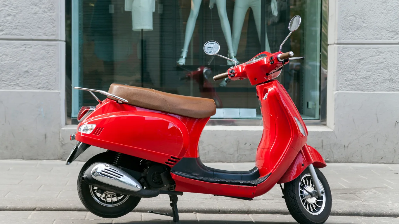 Red scooter with a tan seat parked on a sidewalk in front of a storefront window.