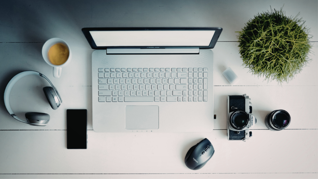 Top-down view of a laptop on a white desk with coffee, headphones, smartphone, mouse, camera and plant nearby.