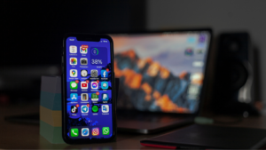 Smartphone with app icons on the home screen on a desk, a blurred laptop in the background.