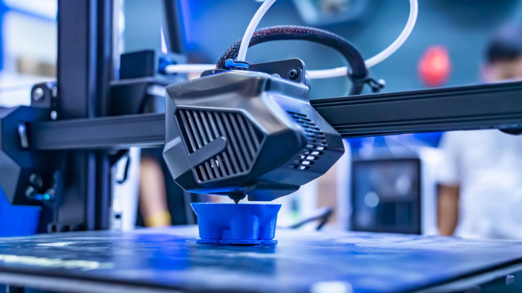 3D printer nozzle extruding blue plastic onto a blue cup-shaped object on the build plate, in a manufacturing setup.