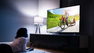 Person lying on the floor watching a large TV that shows a father biking with a child in a trailer along a sunny rural road